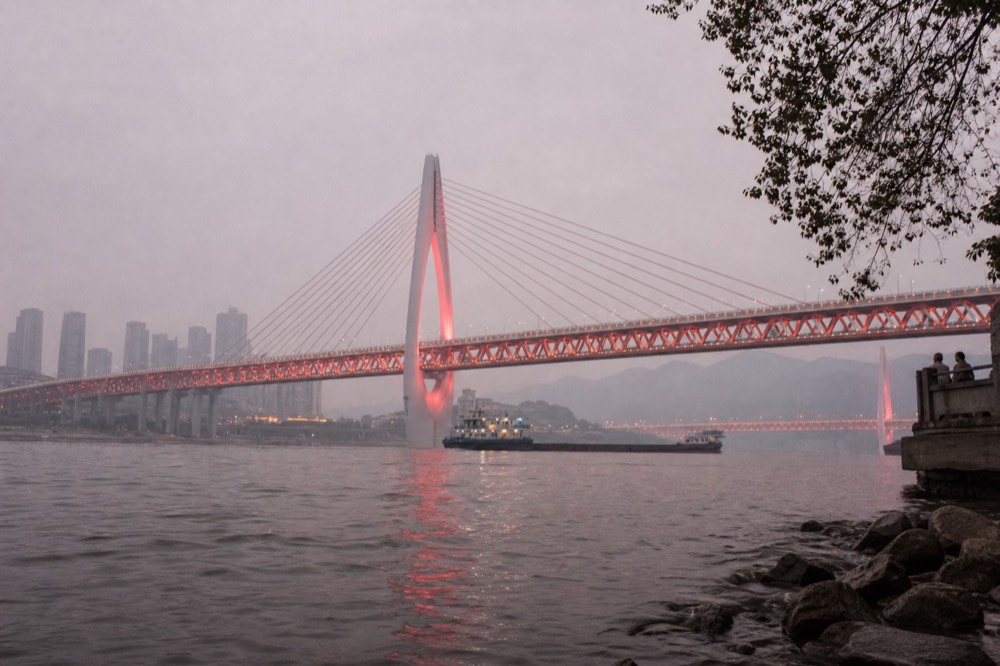 A night-time riverside road with decorative lighting and a lit cable-stay bridge spanning dark water