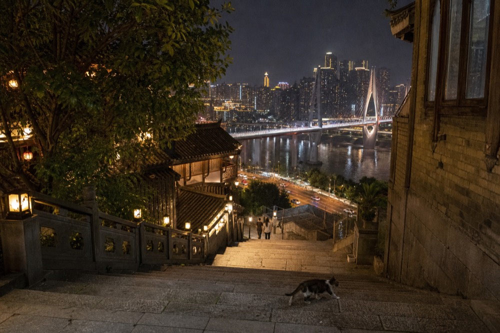 A lit urban bridge and tower skyline seen at night from a riverside promenade with decorative lamp posts