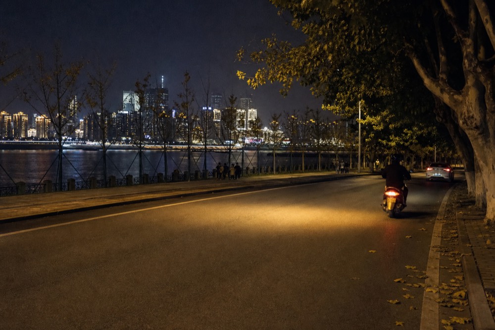 A motorcycle headlight glowing on a dark riverside road with illuminated high-rise towers across the water