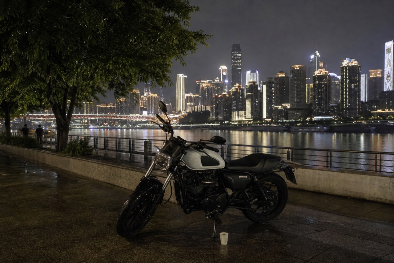 A parked motorcycle on a riverside road at night, with an illuminated city skyline and bridge reflected across the water