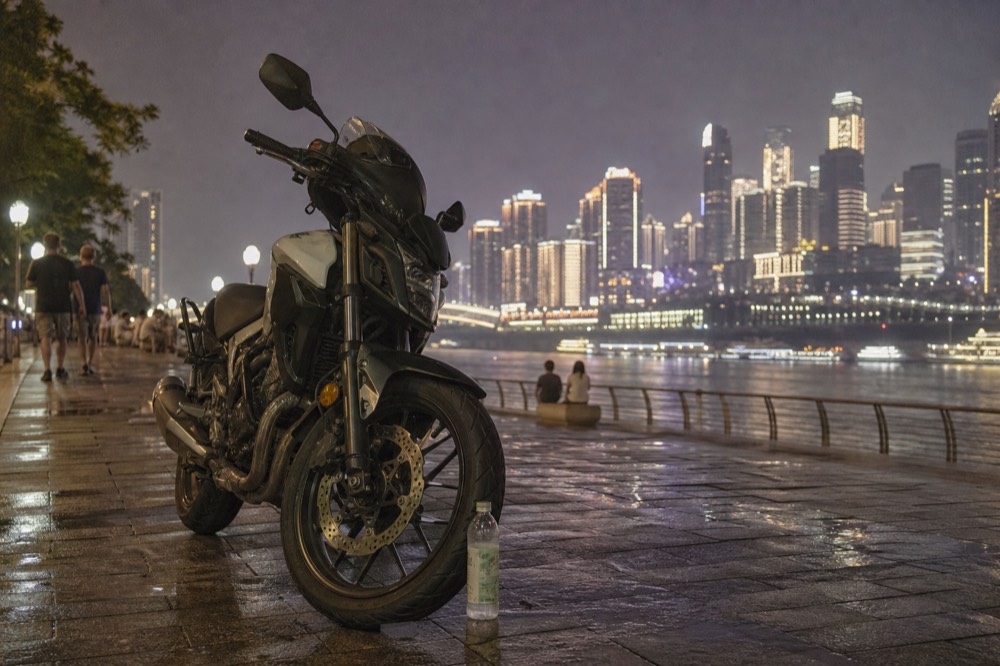 A parked motorcycle on a riverside promenade at night with lit towers across the water