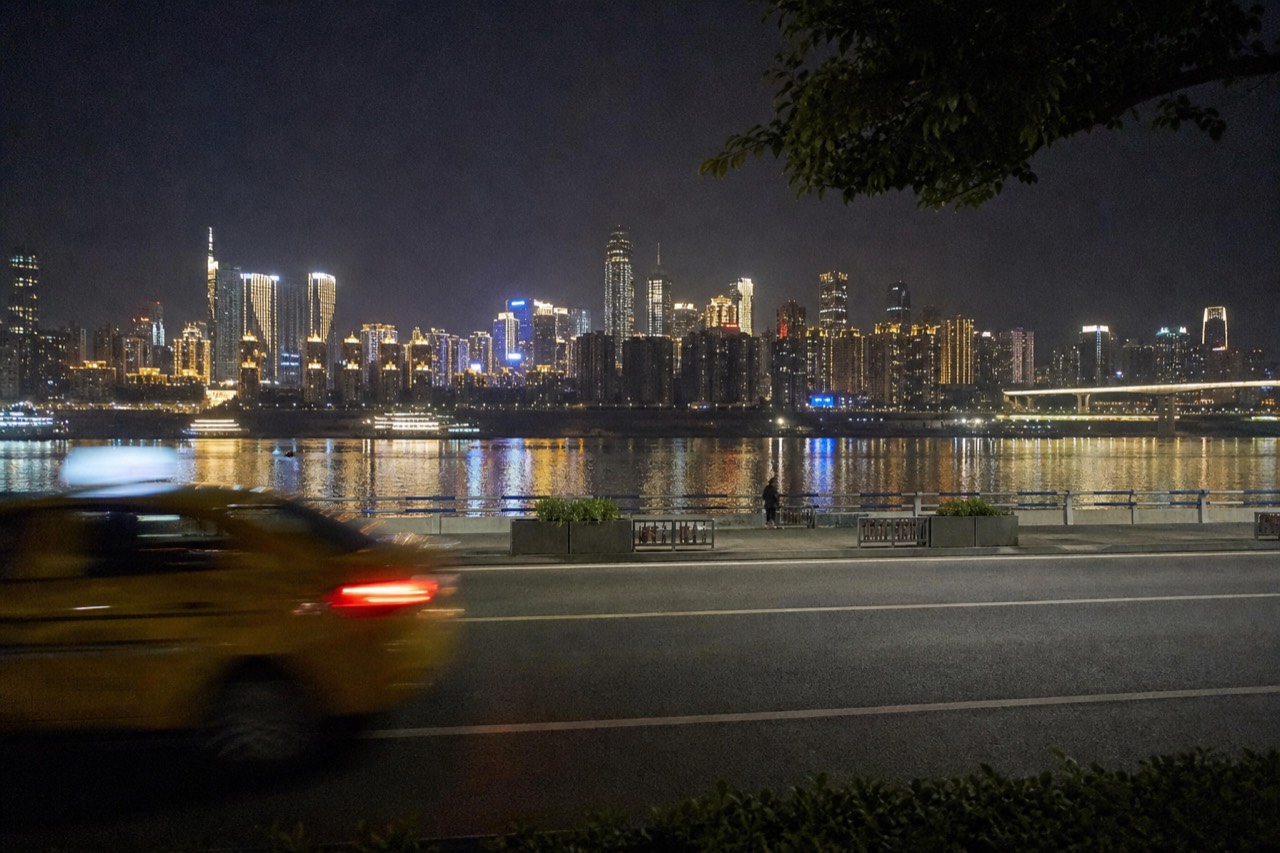 Illuminated city skyline reflected across a wide river at night with a riverside road in the foreground