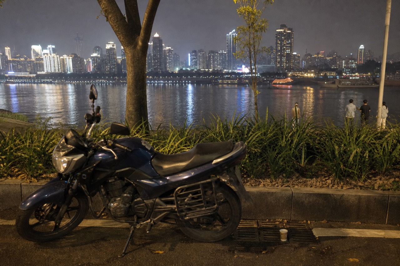 A parked motorcycle on a riverside road with a lit city skyline across the water at night