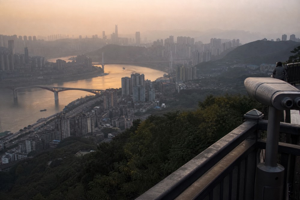 A wide elevated view over dense city rooftops and towers descending toward a river junction at dusk