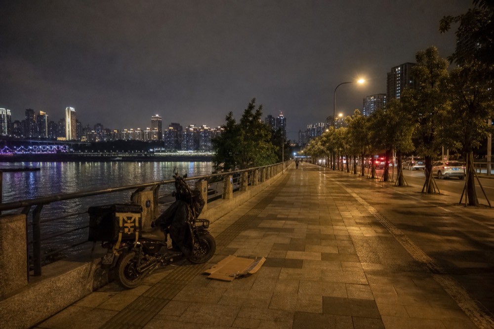 Long riverside road at night with glowing lamp posts and towers rising on the far bank of the river