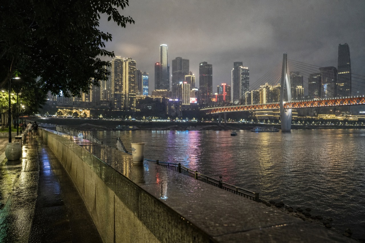 Neon-lit high-rise skyline above a dark river at night, with a lit bridge crossing the water