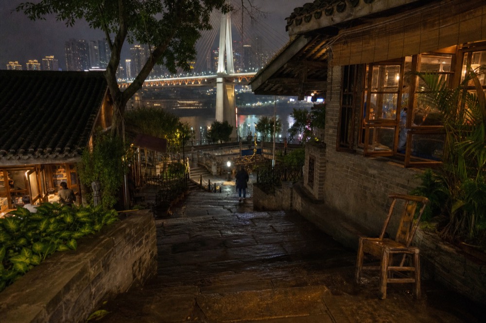 A riverside street at night with neon signs, a lit bridge spanning the water, and towers on the far bank