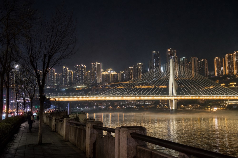 A city skyline of illuminated towers above a dark river, with a cable-span bridge lit in the foreground