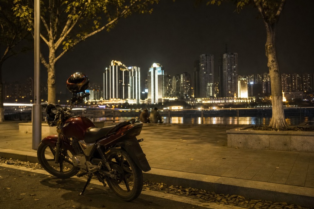 A parked motorcycle in warm street light on a riverside promenade, with illuminated buildings across the water