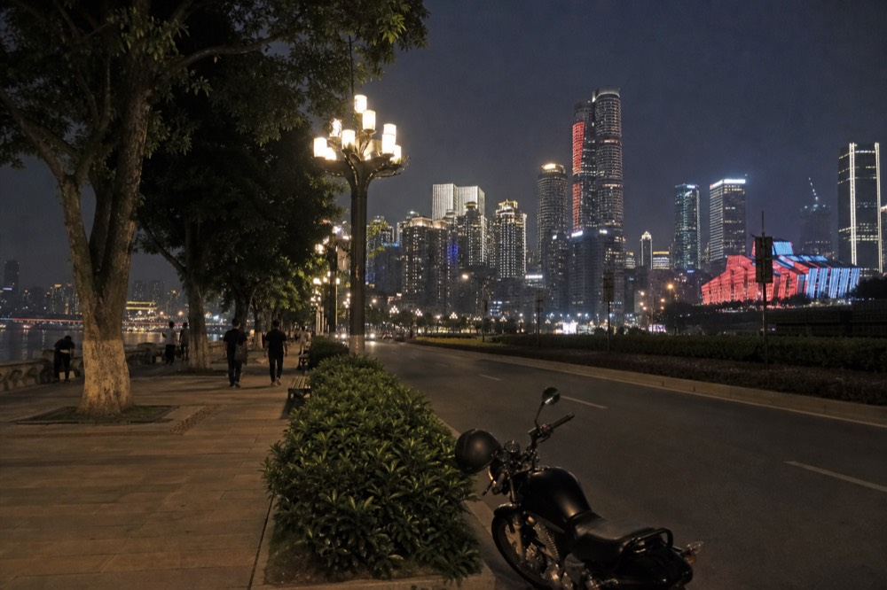 A riverside road at night with decorative lighting along the promenade and city towers across the water