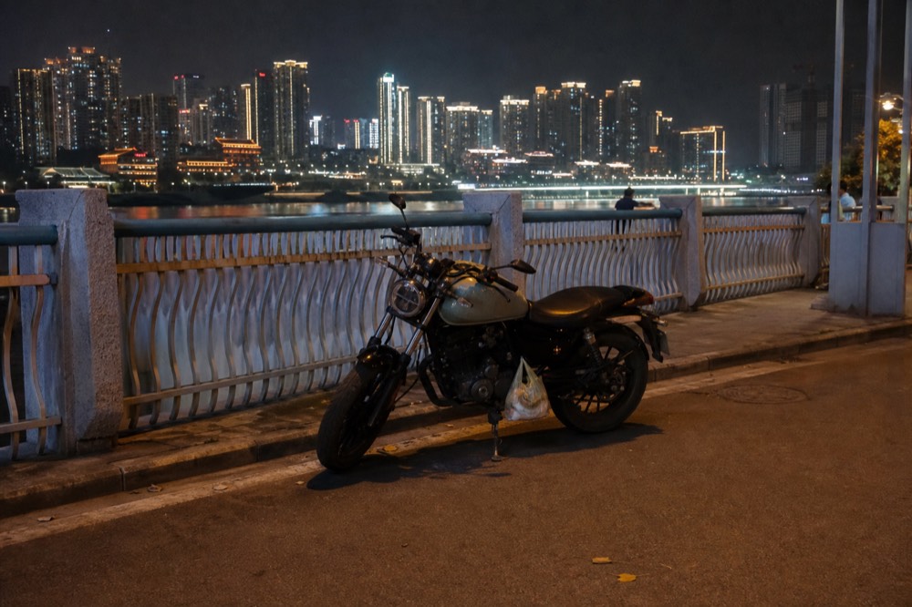 A parked motorcycle beside a riverside promenade with illuminated towers across the river at night