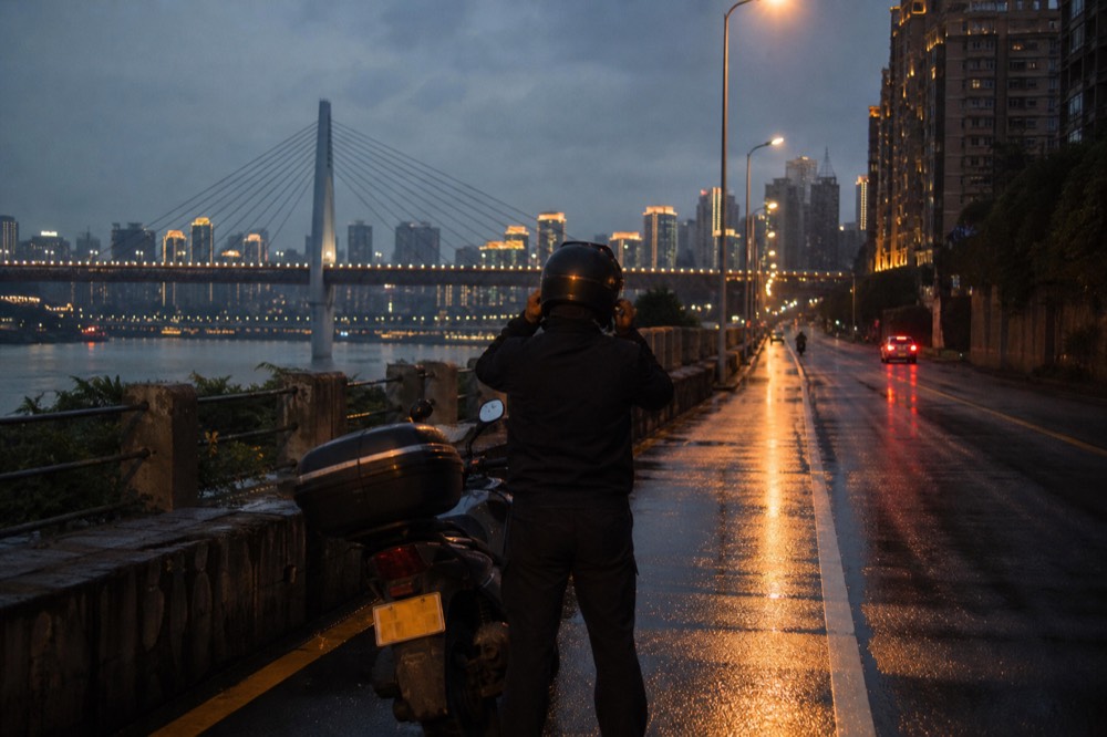 A rider adjusting a helmet beside a motorcycle on a lit riverside street