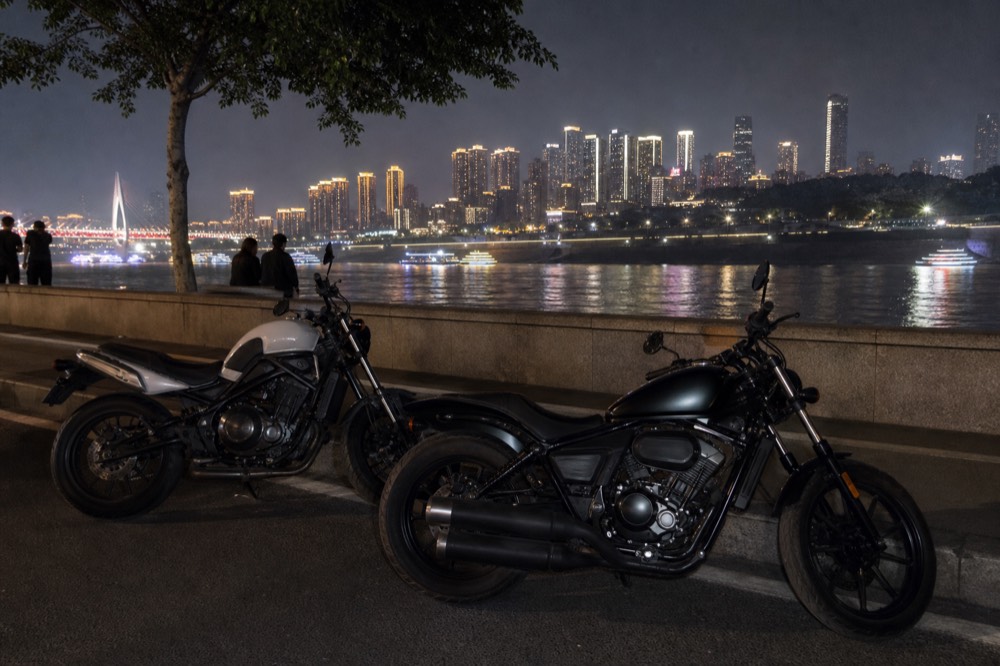 Two motorcycles parked side by side along the low river wall on Nanbin Road at night, the lit Chongqing skyline across the water behind them