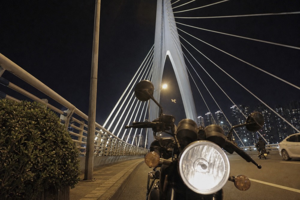 A motorcycle headlight and illuminated bridge cables against a dark sky