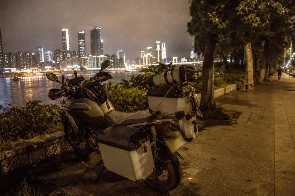 A parked motorcycle with camera equipment strapped to its panniers on a night street, the lit skyline across the river behind