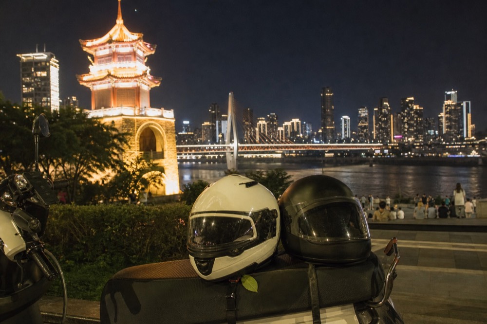 Two helmets resting on a motorcycle seat with a lit bridge and towers across the river at night