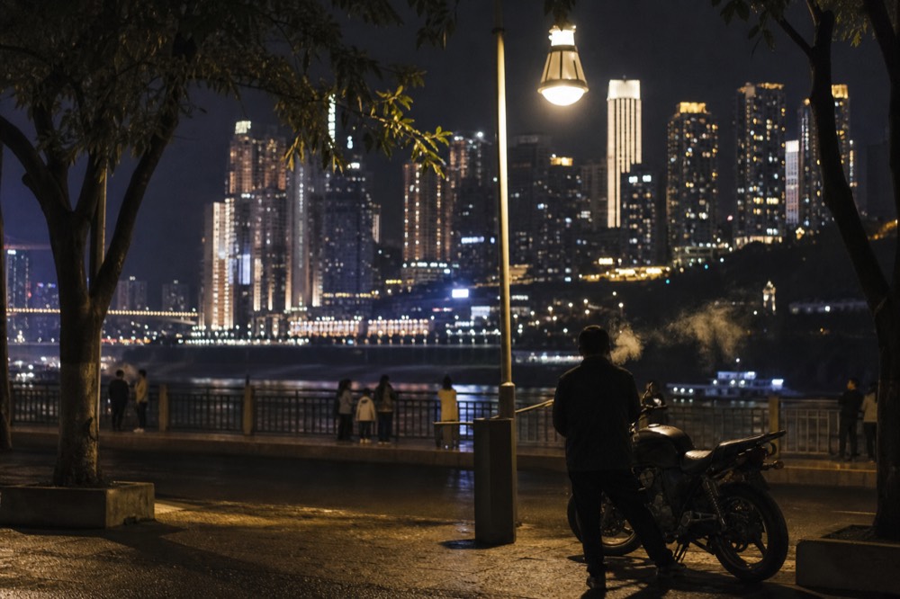 Riverside promenade at night with lit city towers visible across the water