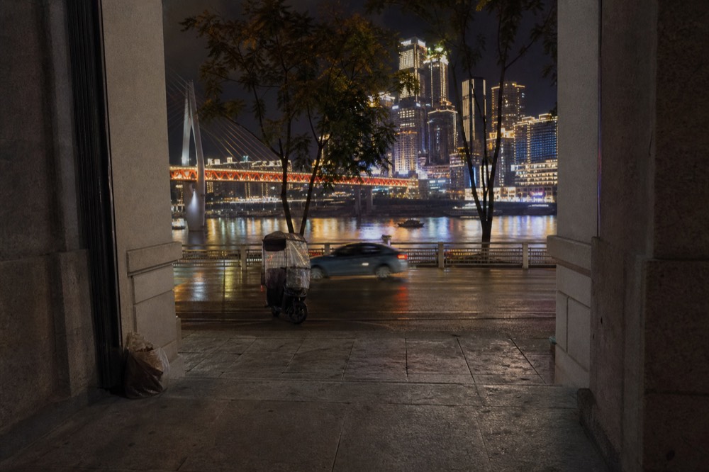 Lit riverside road at night with towers and a bridge visible across the river