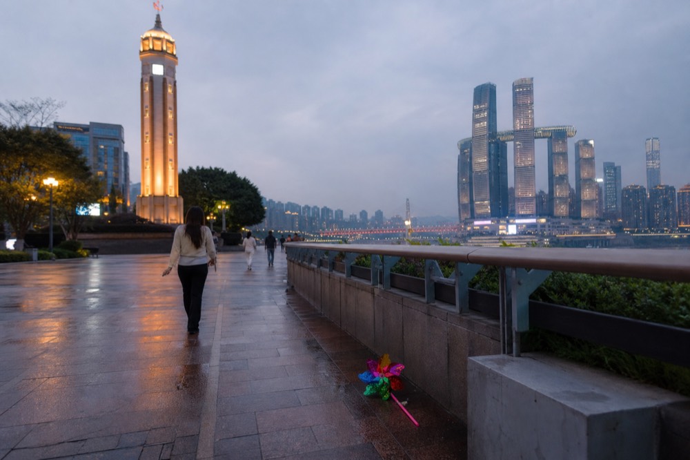 Riverside promenade at night with people walking and lit towers across the river