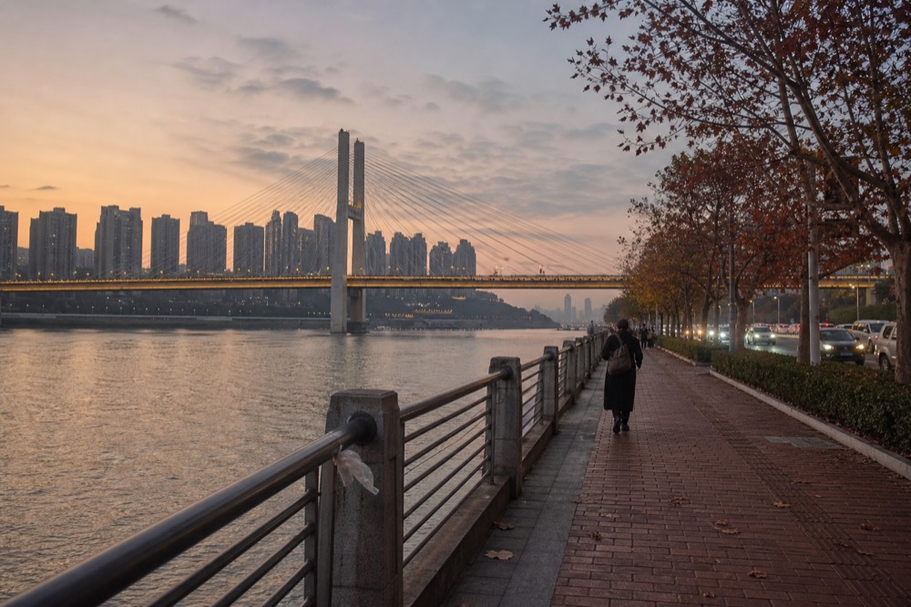Riverside promenade at dusk with a long lit bridge spanning a wide river toward glowing high-rise towers.