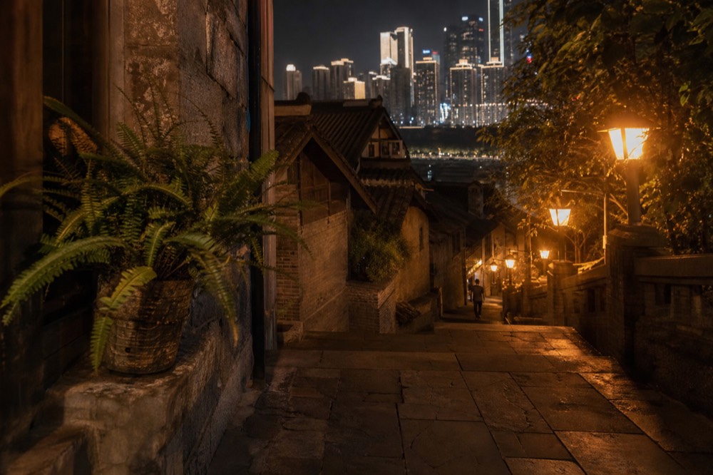Old stone-pedestrian street on a hillside at night, lit by warm lanterns with modern towers glowing behind the rooftops.