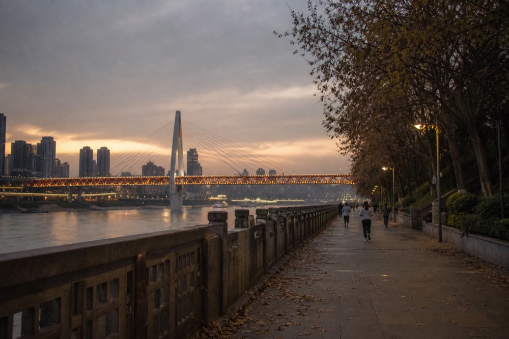 Riverside boulevard at dusk seen from a south bank promenade with a lit bridge spanning the river beyond