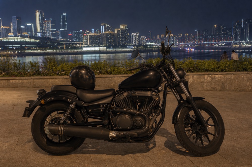 Motorcycle parked on a riverside promenade at night with city skyline lights across the water