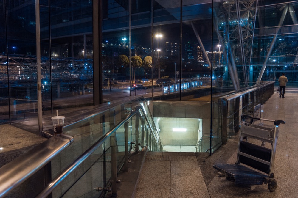 Glass-and-steel concourse at night, softly lit, with blurred figures passing through