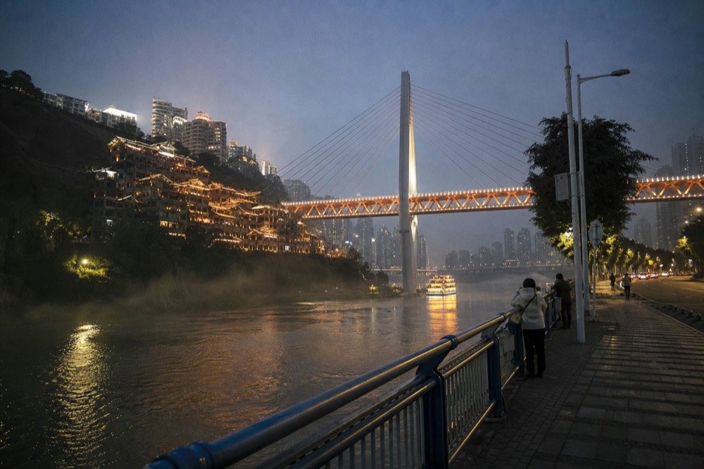 Lit cable-stayed bridge spanning a river above an illuminated stilt-house complex at night