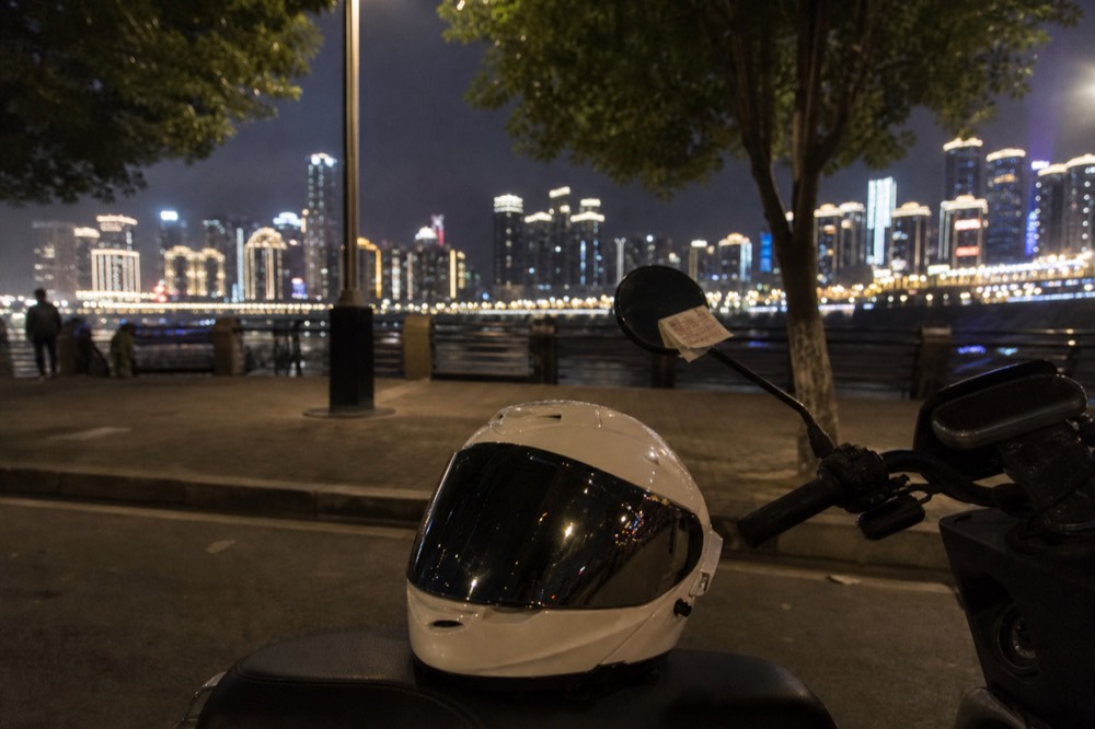 A motorcycle helmet resting on the seat of a parked bike with the city lights of Chongqing in the background