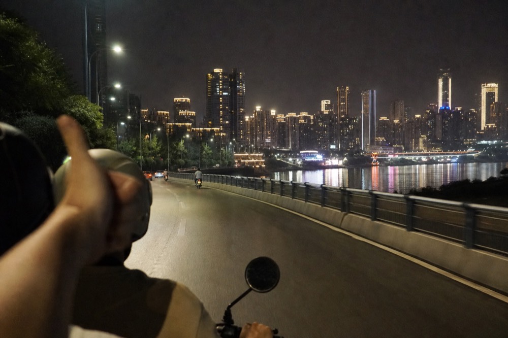 A passenger on a motorcycle giving a thumbs-up to the camera, with the rider in front and city lights behind