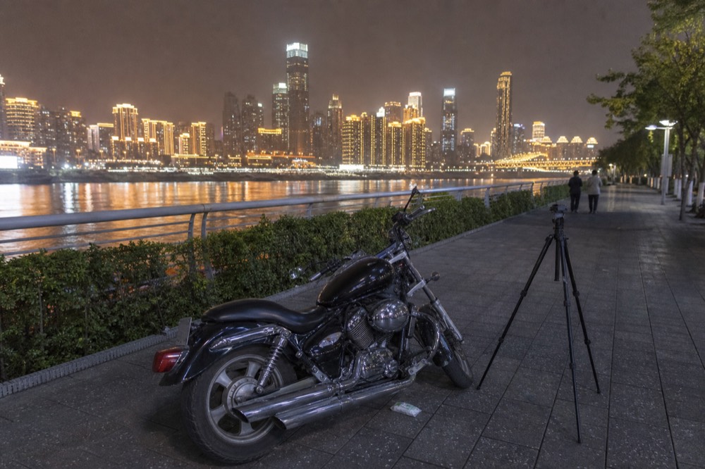 A parked motorcycle on a riverside promenade with lit towers across the water at night