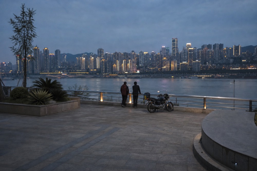 A riverside plaza on Nanbin Road at dusk, two figures at the rail with the Chongqing skyline across the Yangtze