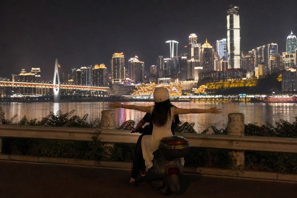 A motorcycle passenger posing with arms outstretched during a night shoot along Nanbin Road with the lit skyline behind