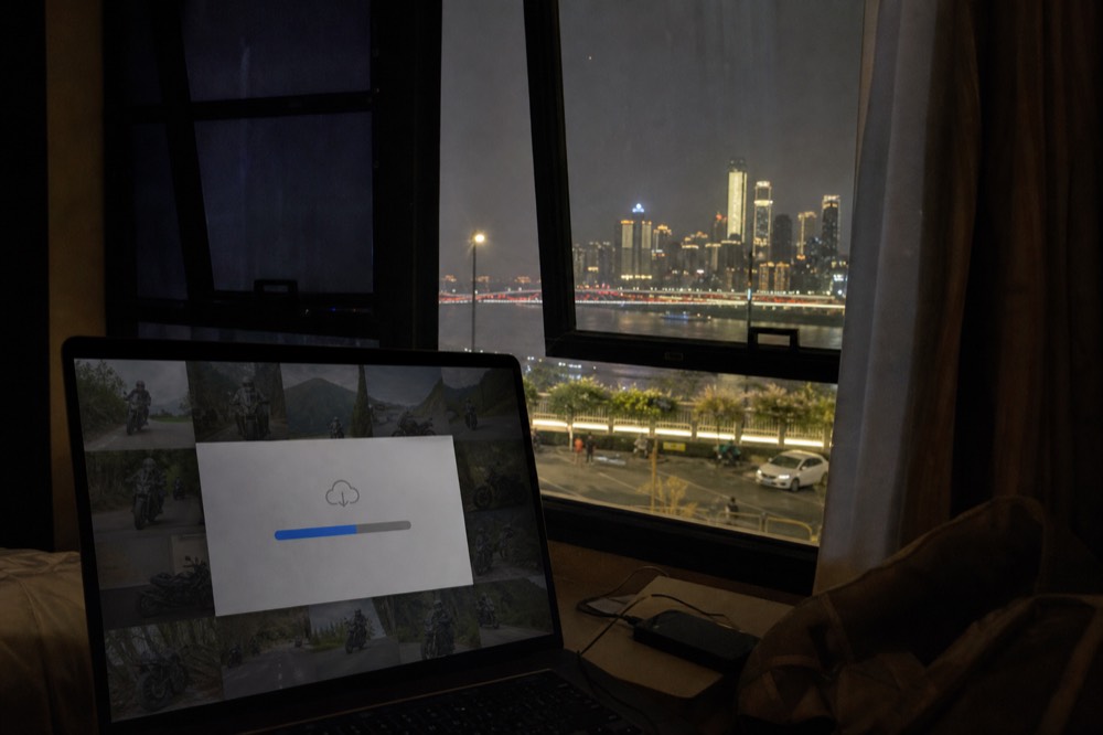 A laptop screen showing a folder of motorcycle photos downloading from a cloud link, the lit Chongqing skyline through the window behind