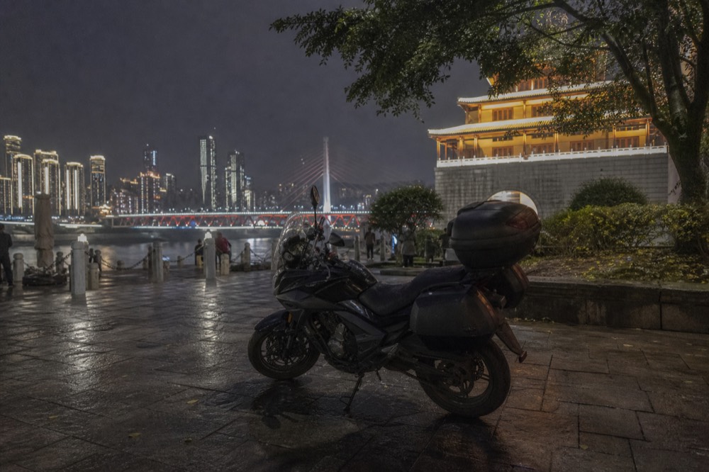 A motorcycle parked on a riverside promenade at night with city lights across the water