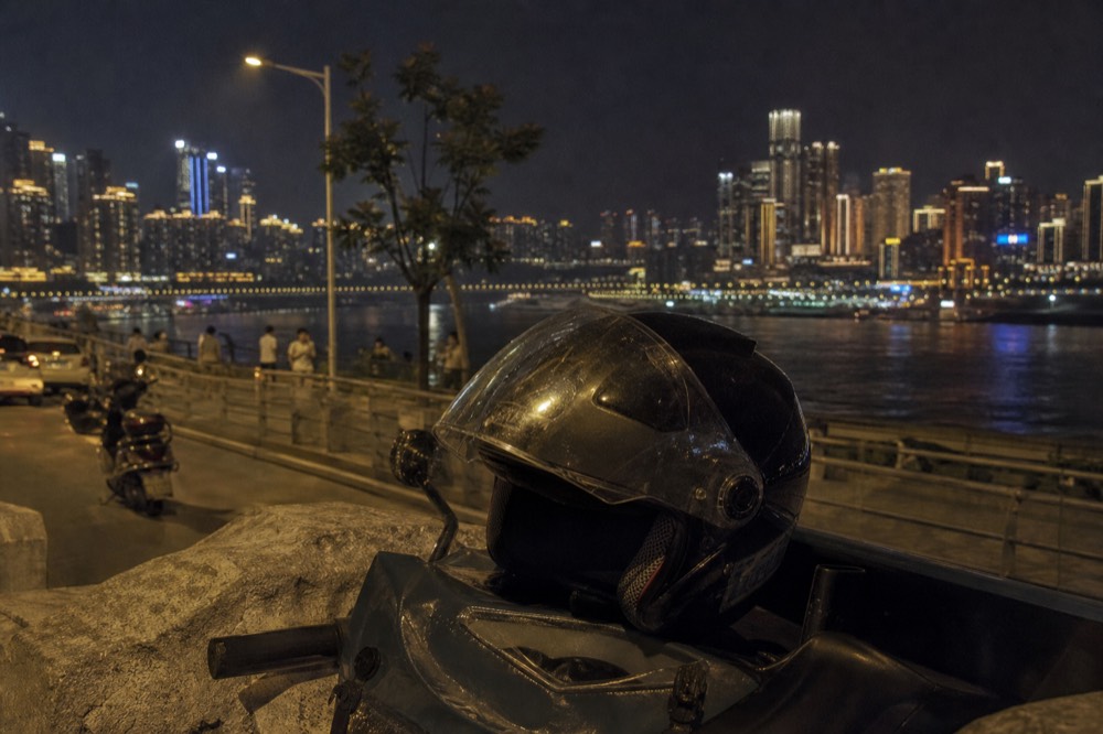 A motorcycle helmet resting on a bag on the river wall with the lit city across the water behind