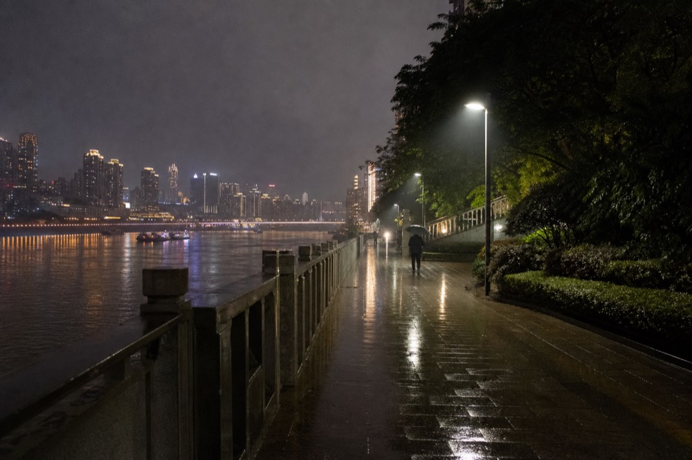 Riverside walkway at night with lamp posts and the glowing skyline of the opposite bank beyond the water