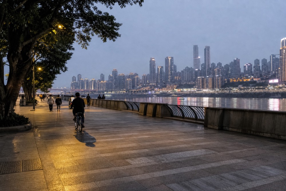 A riverside promenade at dusk with street lamps lit and city towers glowing across the water