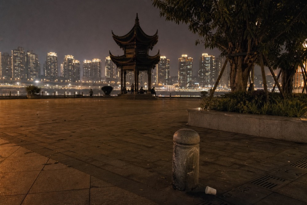 A plaza by a river at night with warm lamps and distant illuminated high-rises under a dark sky