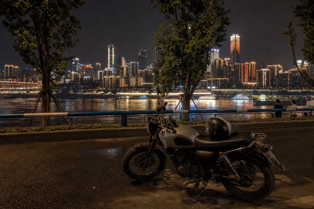 A motorcycle parked on a riverside road at night with a brightly lit skyline of towers behind it under a black sky