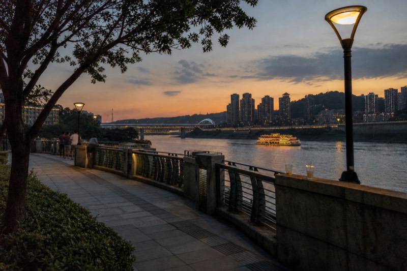 The riverside promenade in Chongqing on a warm evening, a handful of people at the railings watching ferries cross the lit stretch of the Yangtze.