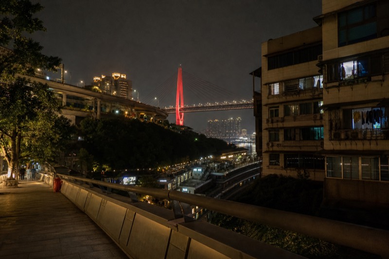 Chongqing's tiered hillside form at night, with buildings and roadways stacked at several levels above the black water of the Yangtze and the lit bridge beyond.